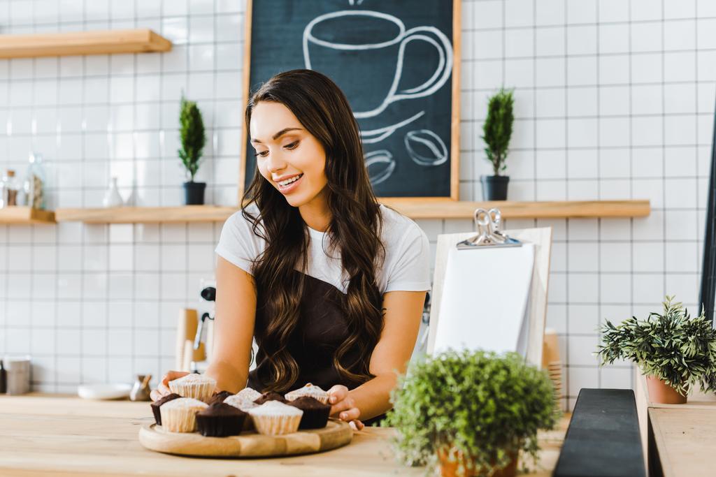 привлекательная официантка, стоящая за барной стойкой и держащая поднос с кексами в кофейне
 - Фото, изображение
