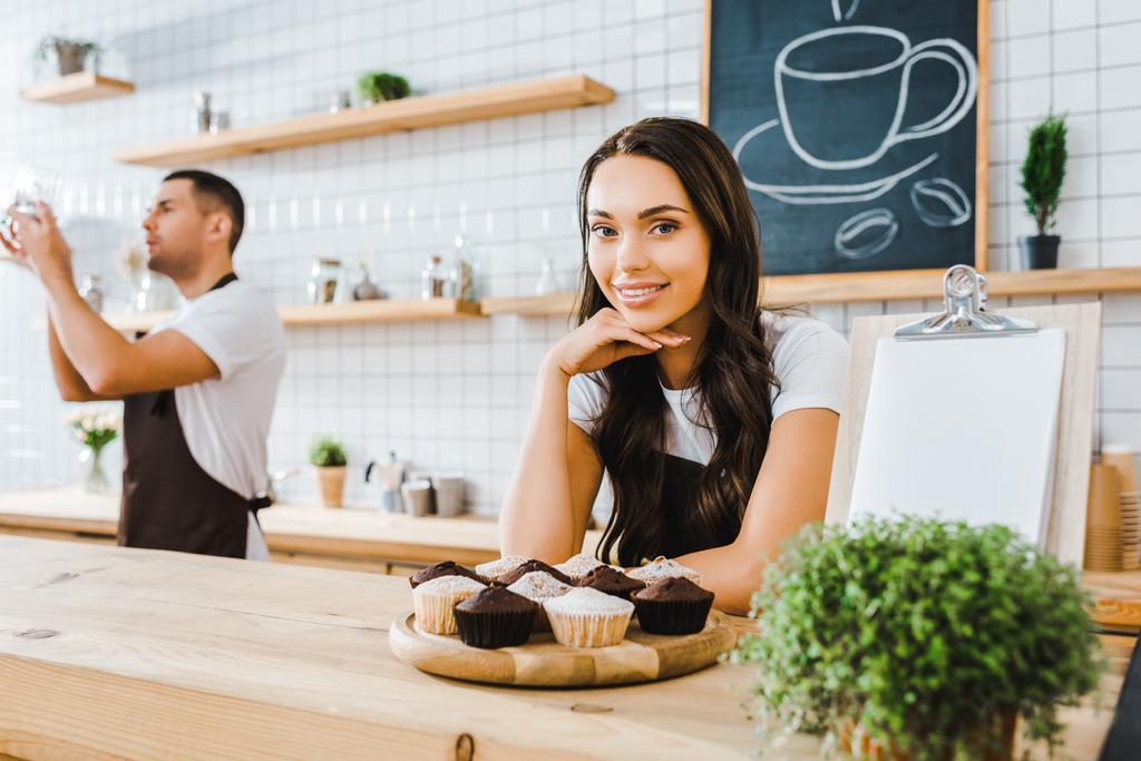 Ελκυστική μελαχρινή σερβιτόρα στέκεται πίσω από μπαρ με cupcakes σε ξύλινη θήκη barista πανουργία κρατώντας το κύπελλο στο καφενείο - Φωτογραφία, εικόνα