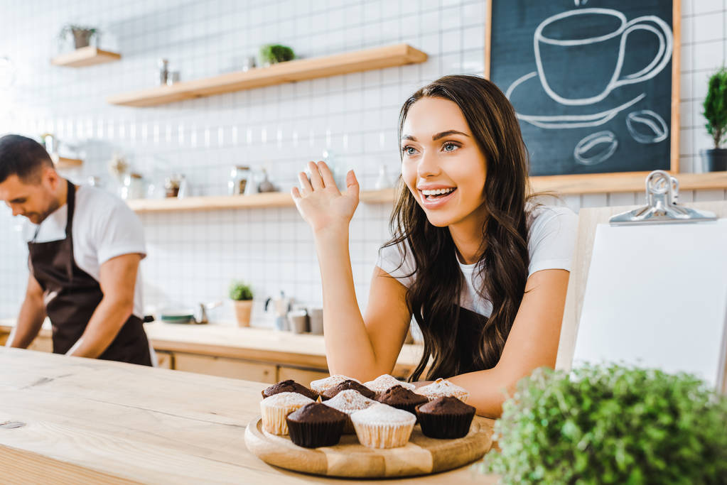 вибірковий фокус привабливої брюнетки офіціантки, що стоїть за барною стійкою з кексами і махаючим касиром, що працює в кав'ярні
 - Фото, зображення
