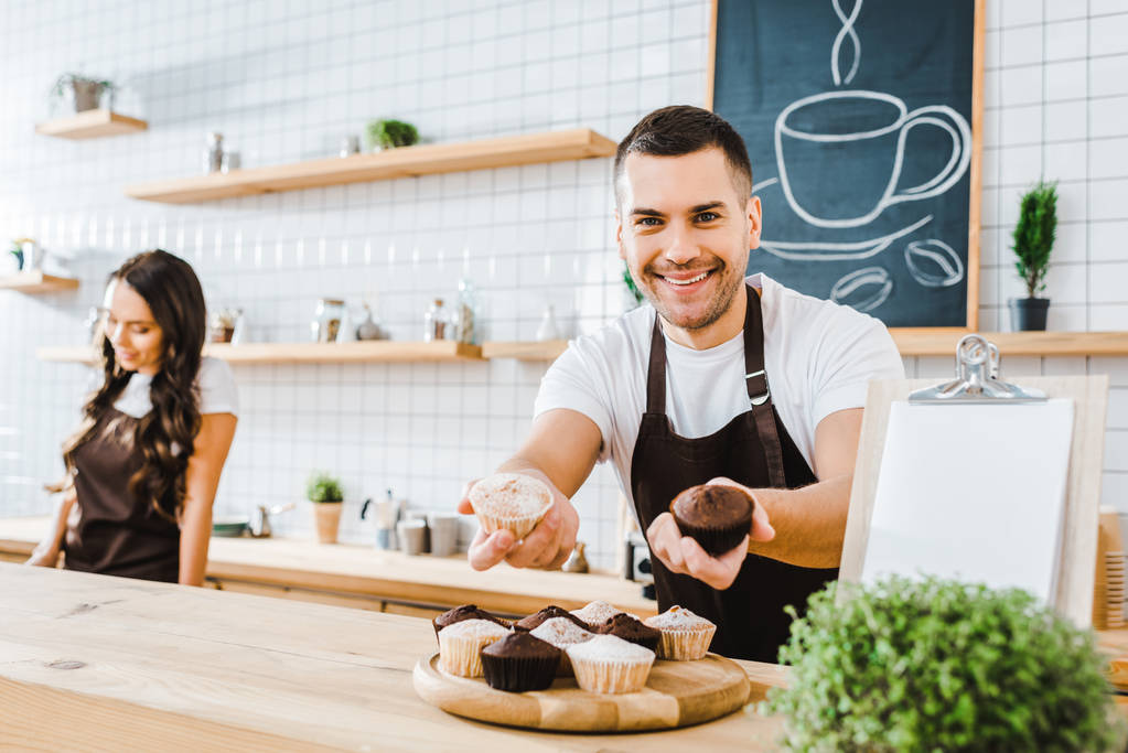 selectieve aandacht van knappe caissière in schort geven cupcakes wile barista werken in koffiehuis - Foto, afbeelding