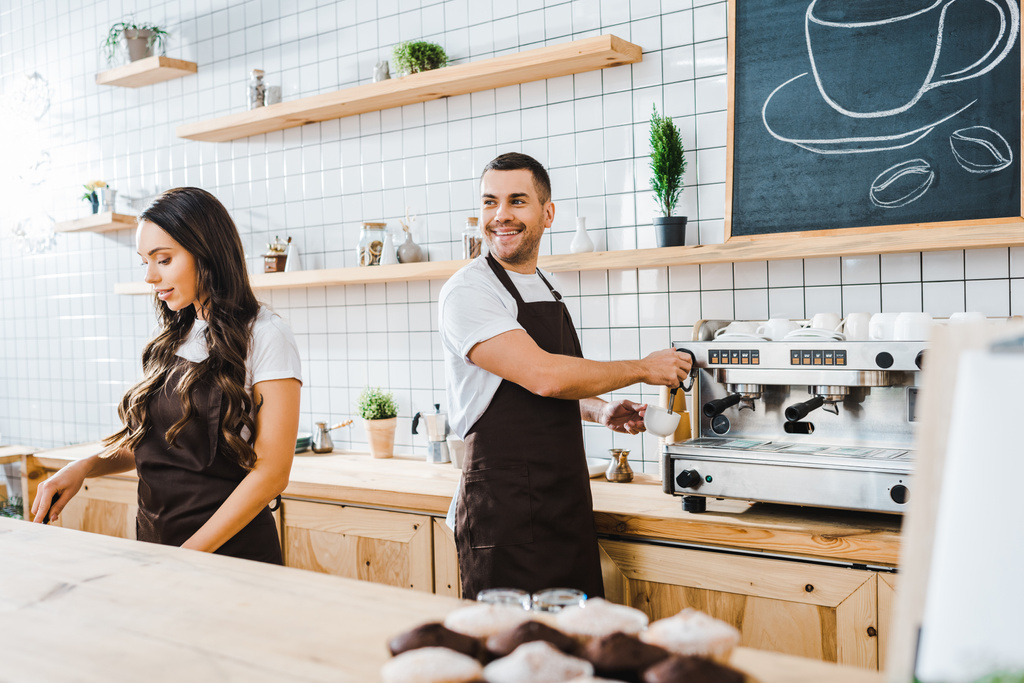 barista haciendo café y sonriendo astuto cajero atractivo trabajando detrás del mostrador de la barra en la cafetería
 - Foto, Imagen