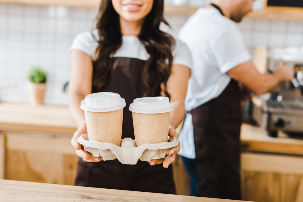 обрезанный вид брюнетки кассир стоя и держа бумажные чашки, коварный бариста работает в кофейне
 - Фото, изображение