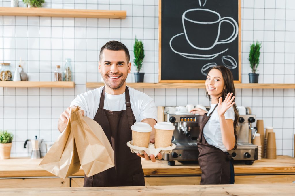 handsome cashier holding paper cups and bags wile attractive brunette barista wavingin in coffee house - Photo, Image