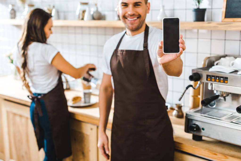 Hübsche Kassiererin zeigt Smartphone mit leerem Bildschirm: Barista arbeitet im Kaffeehaus - Foto, Bild