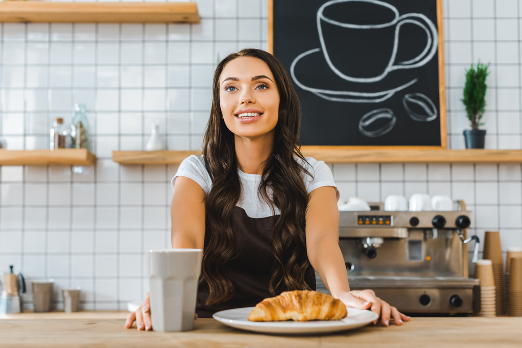 attractive cashier standing behind bar counter with cup, plate and croissant in coffee house - Photo, Image