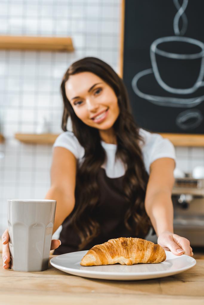 selective focus of attractive brunette cashier standing behind bar counter with cup, plate and croissant in coffee house - Photo, Image