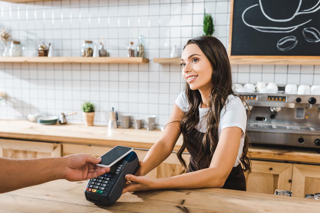 attraktive Kassiererin, die in brauner Schürze neben der Theke steht und im Kaffeehaus mit dem Smartphone bezahlt - Foto, Bild