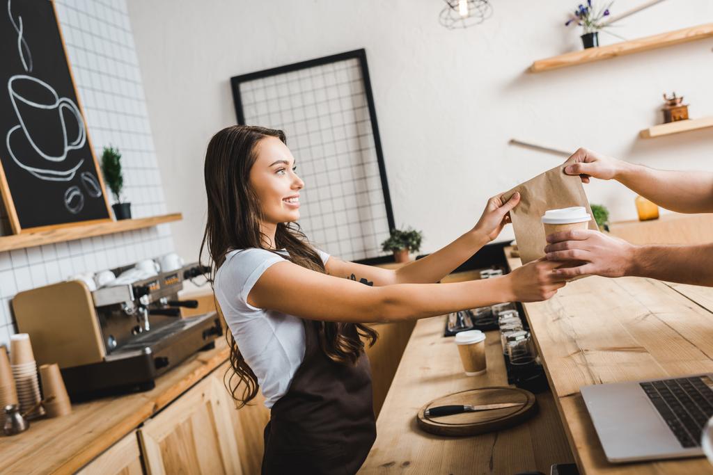 aantrekkelijke Kassier in bruin schort geven papier cup en tas aan de mens in koffiehuis - Foto, afbeelding