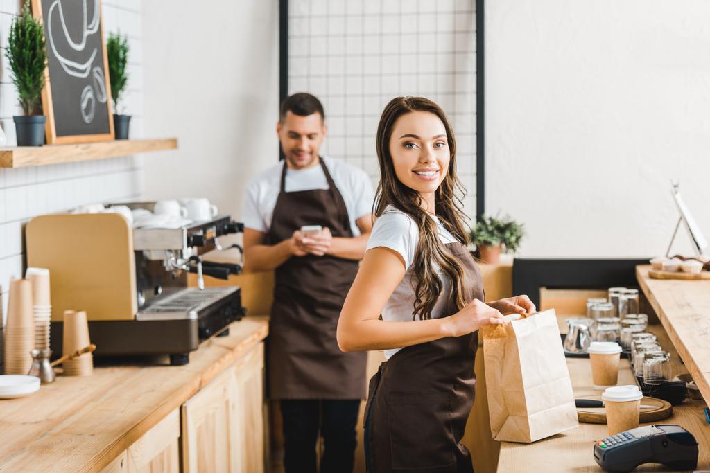 cashier with paper bag smiling and barista with smartphone standing behind bar counter in coffee house - Photo, Image
