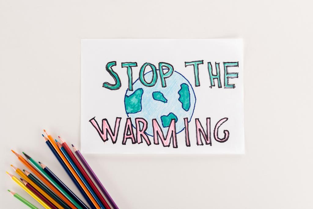 card with earth symbol and "stop warming" inscription, and multicolored pencils on white background - Photo, Image