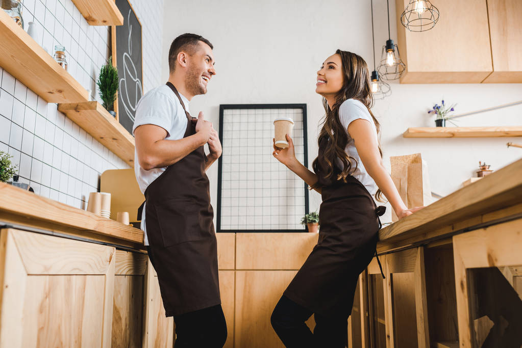 Pokladník drží pohárek a povídání s barista v hnědá zástěra poblíž dřevěný bar pult v kavárně - Fotografie, Obrázek