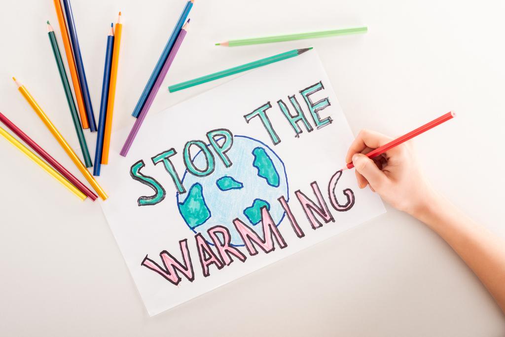 bijgesneden beeld van vrouw schrijven 'stop de opwarming van de aarde' op kaart met globe teken en veelkleurige potloden op witte achtergrond - Foto, afbeelding