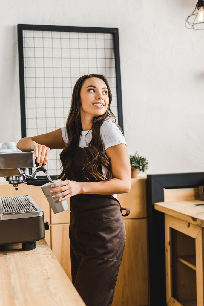 attraktive brünette Barista in brauner Schürze Tasse halten und Kaffee kochen mit Maschine im Kaffeehaus - Foto, Bild