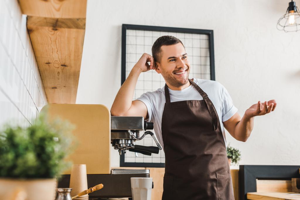 Улыбающийся красивый бариста в коричневом фартуке стоит рядом с кофеваркой в кофейне
 - Фото, изображение
