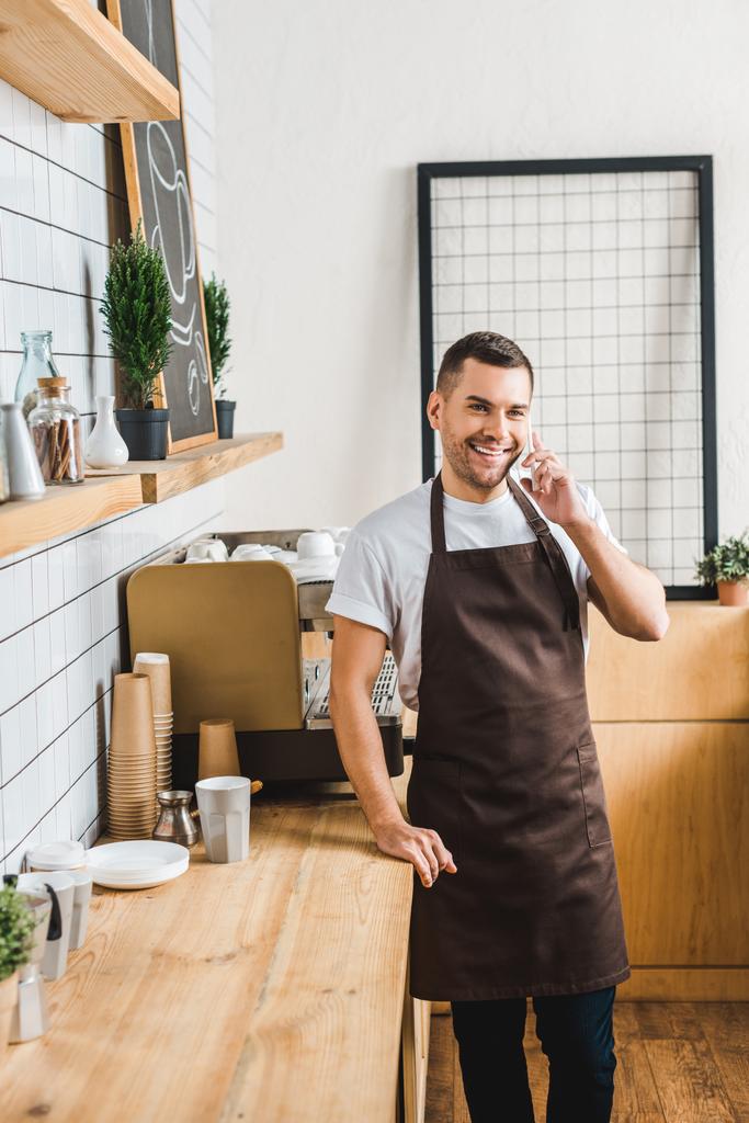 przystojny Kasjera w brązowy fartuch rozmowy na smartfon w pobliżu ekspres do kawy i licznik w coffee house bar - Zdjęcie, obraz