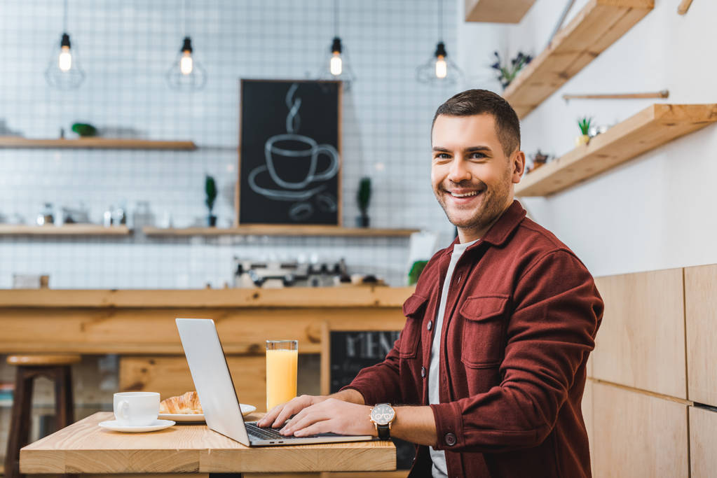 handsome freelancer sitting at table with laptop and smiling in coffee house - Photo, Image