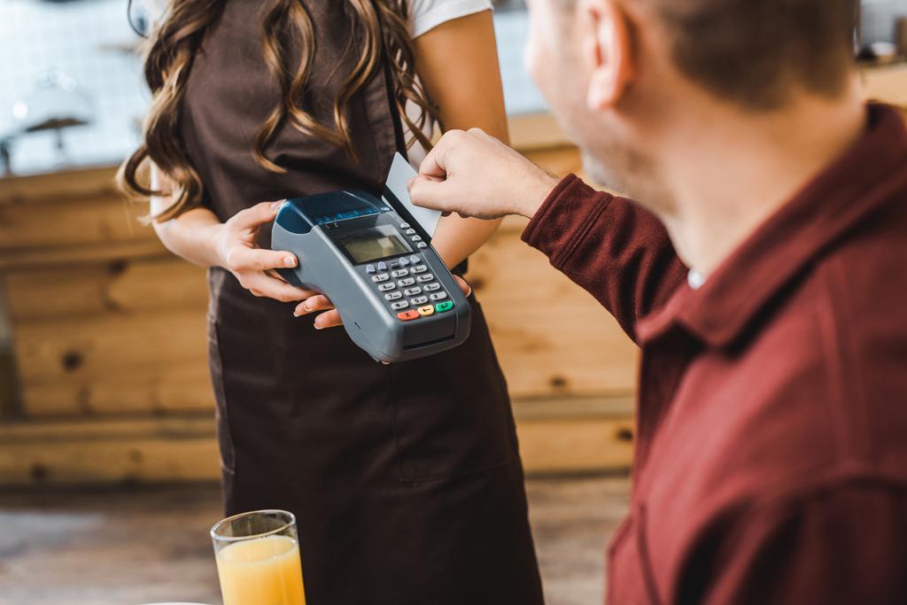 cropped view of waitress holding terminal wile handsome man sitting at table and paying with credit card in coffee house - Photo, Image