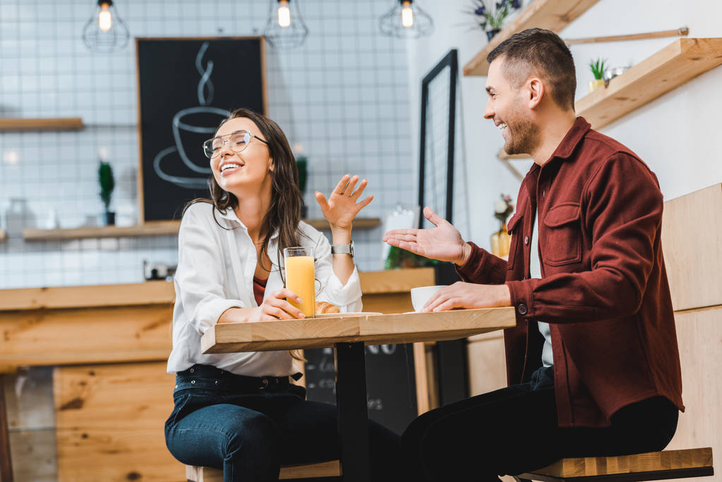 привлекательная брюнетка женщина со стаканом сока и красивый мужчина с чашкой сидя и смеясь за столом в кофейне
 - Фото, изображение