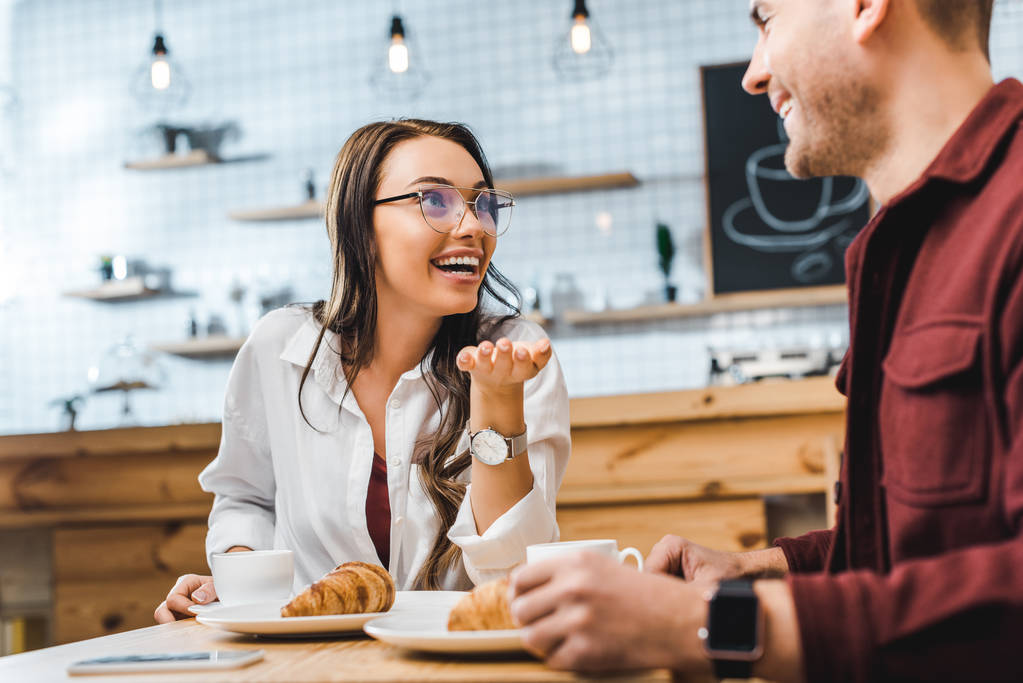 vonzó barna nő és jóképű férfi ül asztal, croissant, bordó ing mosolyogva, és beszél a kávéházban - Fotó, kép