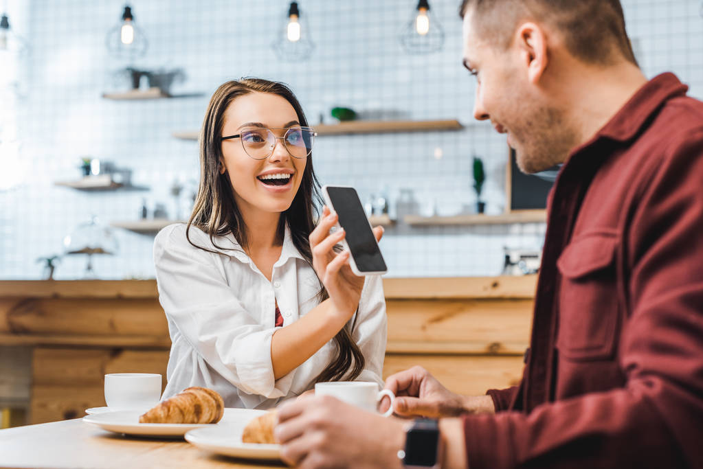 atractiva mujer morena sentada en la mesa y mostrando teléfono inteligente con pantalla en blanco para hombre guapo en camisa de color burdeos en la cafetería
 - Foto, Imagen