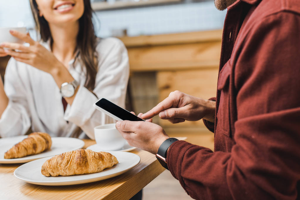 cropped view of man holding smartphone with blank screen wile woman sitting at table and smiling in coffee house - Photo, Image
