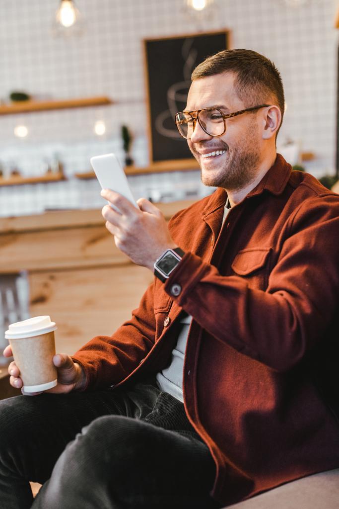 man in burgundy shirt sitting on couch, holding paper cup, looking to smartphone and laughing in coffee house - Photo, Image