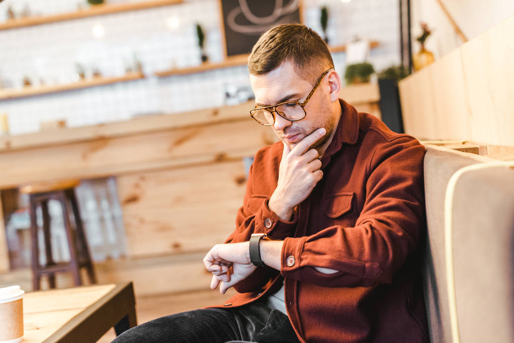 handsome man in burgundy shirt and black jeans sitting on couch and looking to watch with finger in coffee house - Photo, Image