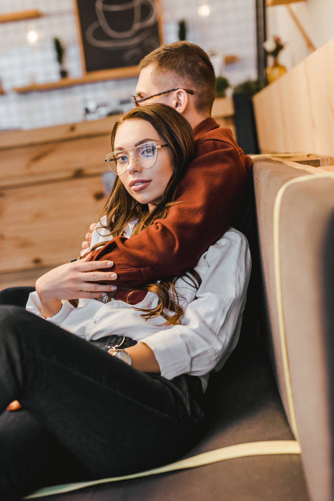 mulher atraente em camisa branca e homem de óculos sentado no sofá e abraçando na casa de café
 - Foto, Imagem