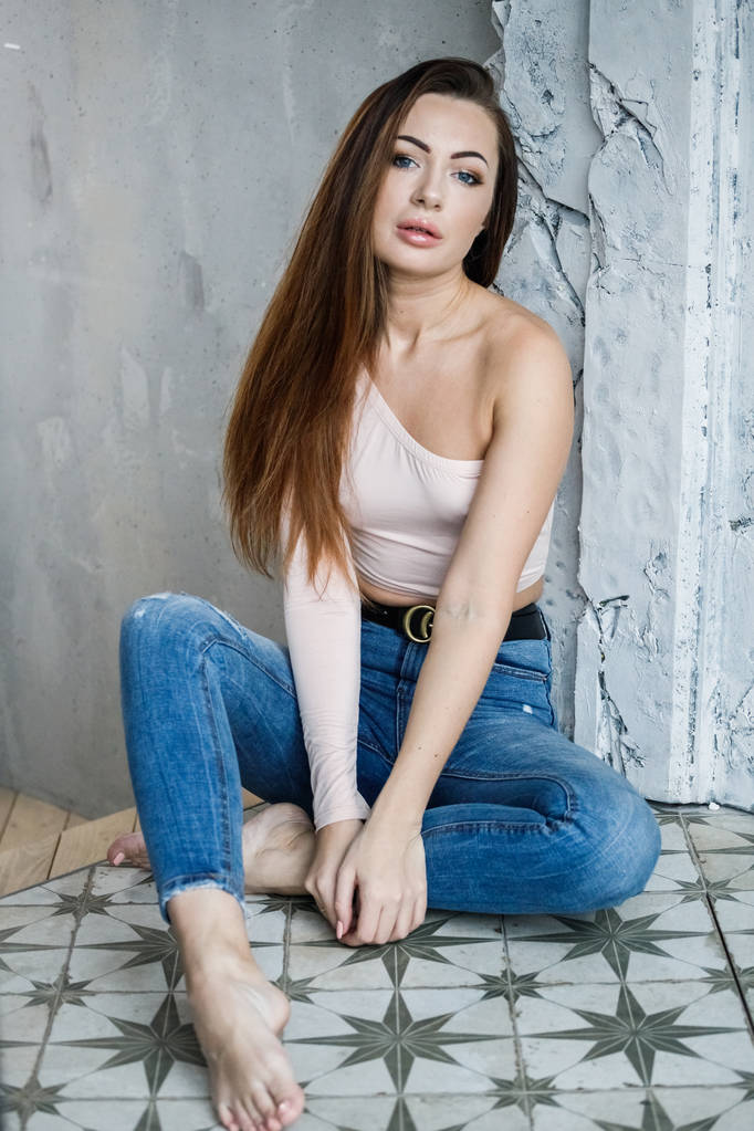 Portrait de belle jeune femme en jean bleu et t-shirt beige. Jeune modèle assis sur le sol près du mur à l'intérieur des appartements de luxe
 - Photo, image