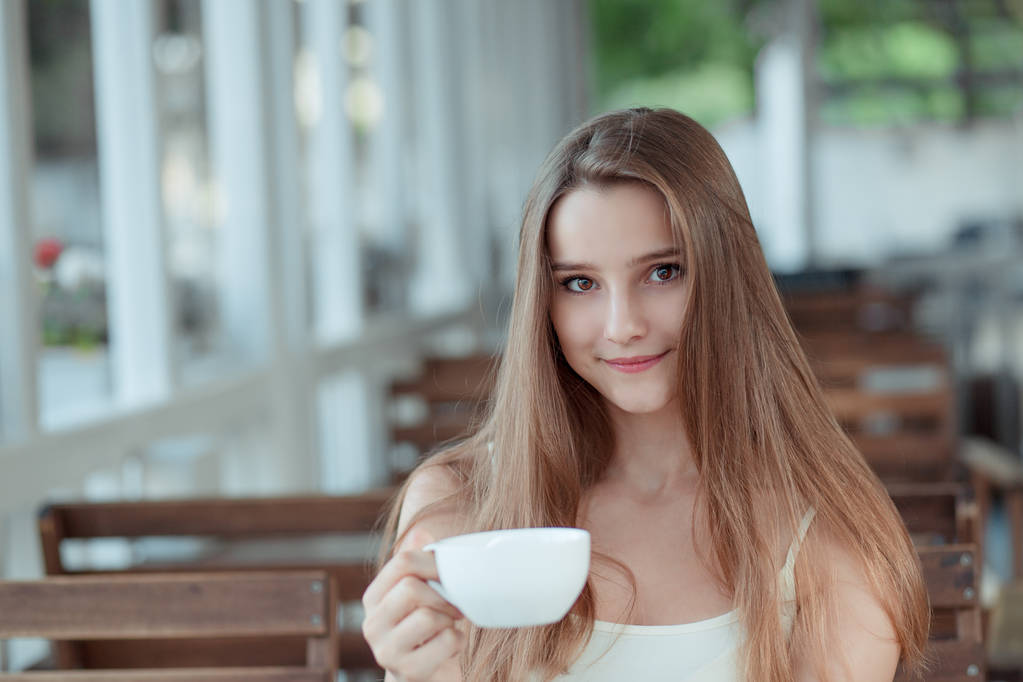 Nuori nainen, teinityttö kahvikupin ääressä katselemassa sinua kameran hymyillessä parvekkeella kahvilassa. Monikulttuurinen malli, sekarotuinen, aasialainen venäläinen tyttö. Vaakasuora. Ihmiset rentouttava käsite
 - Valokuva, kuva