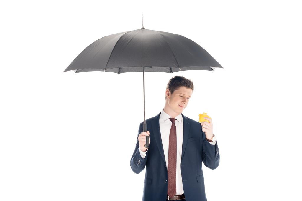 smiling young businessman with umbrella looking at piggy bank isolated on white  - Photo, Image