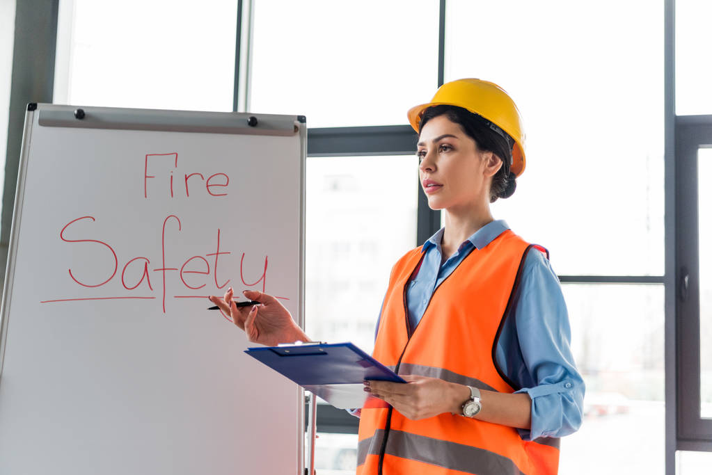Женщина-пожарный в шлеме держит планшет и ручку, стоя рядом с белой доской с надписью пожарной безопасности
 - Фото, изображение