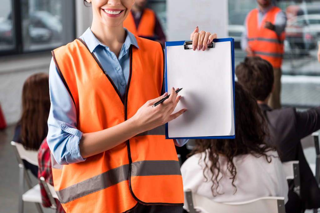 cropped view of cheerful female firefighter pointing at blank clipboard with pen near coworkers giving talk on briefing on background - Photo, Image