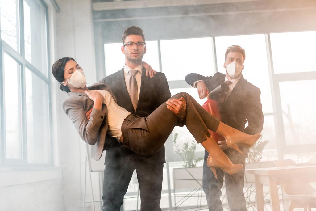 handsome businessman in glasses holding in arms female coworker in mask near colleague holding extinguisher in office with smoke - Photo, Image