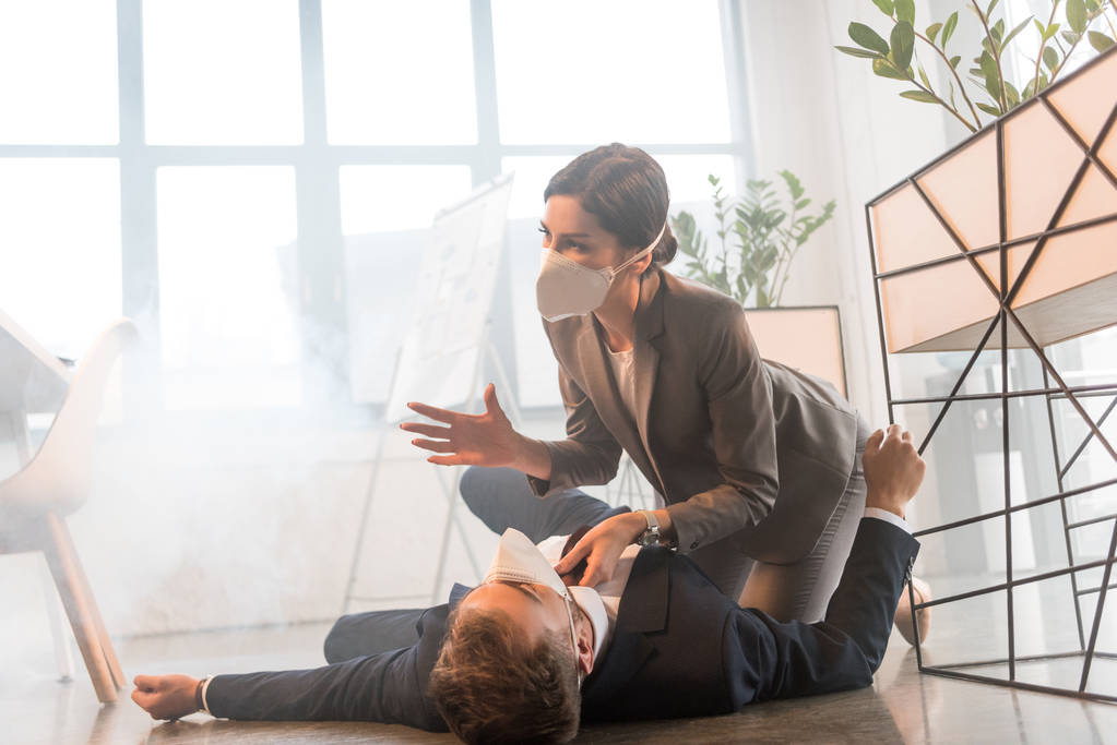przerażona Kobieta w masce siedzi w pobliżu kolegi leżącego na podłodze w biurze z dymu - Zdjęcie, obraz