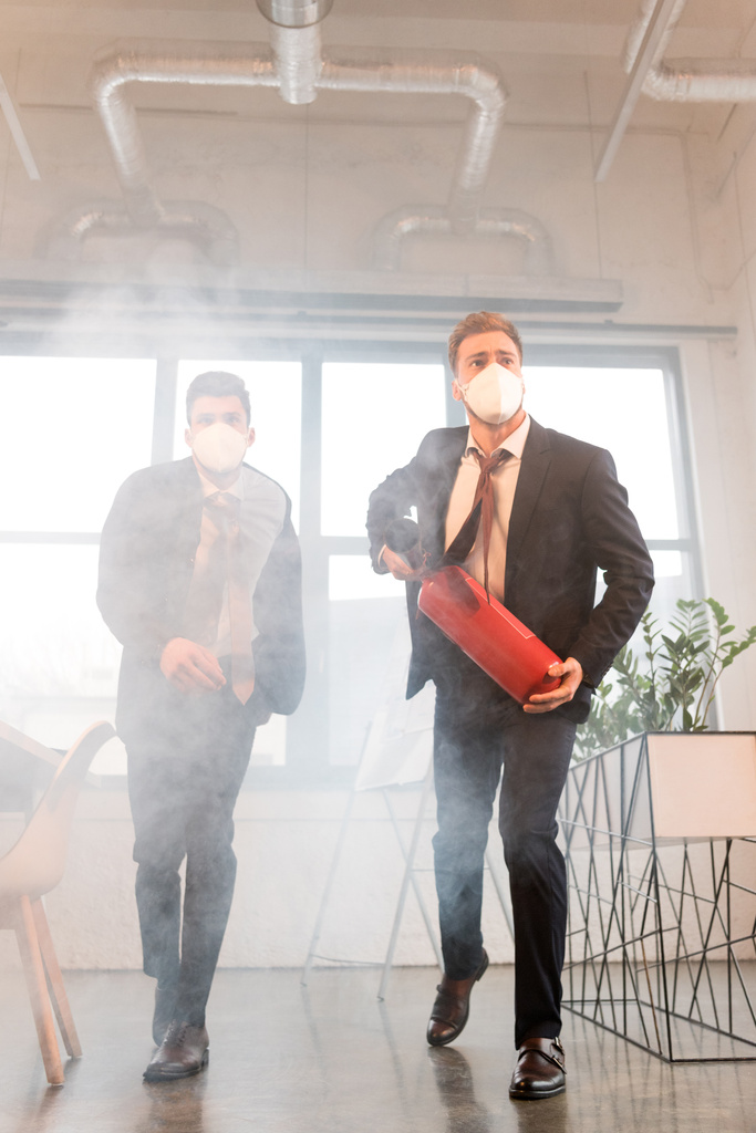 uomo d'affari in maschera che tiene l'estintore vicino al collega mentre cammina in ufficio con il fumo
 - Foto, immagini