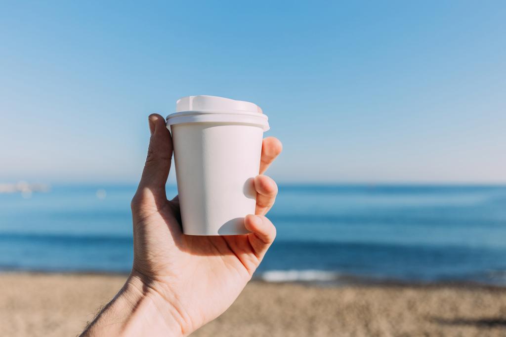 частичный вид человека, держащего бумажную чашку на спокойном синем фоне моря, Барселона, Испания
 - Фото, изображение