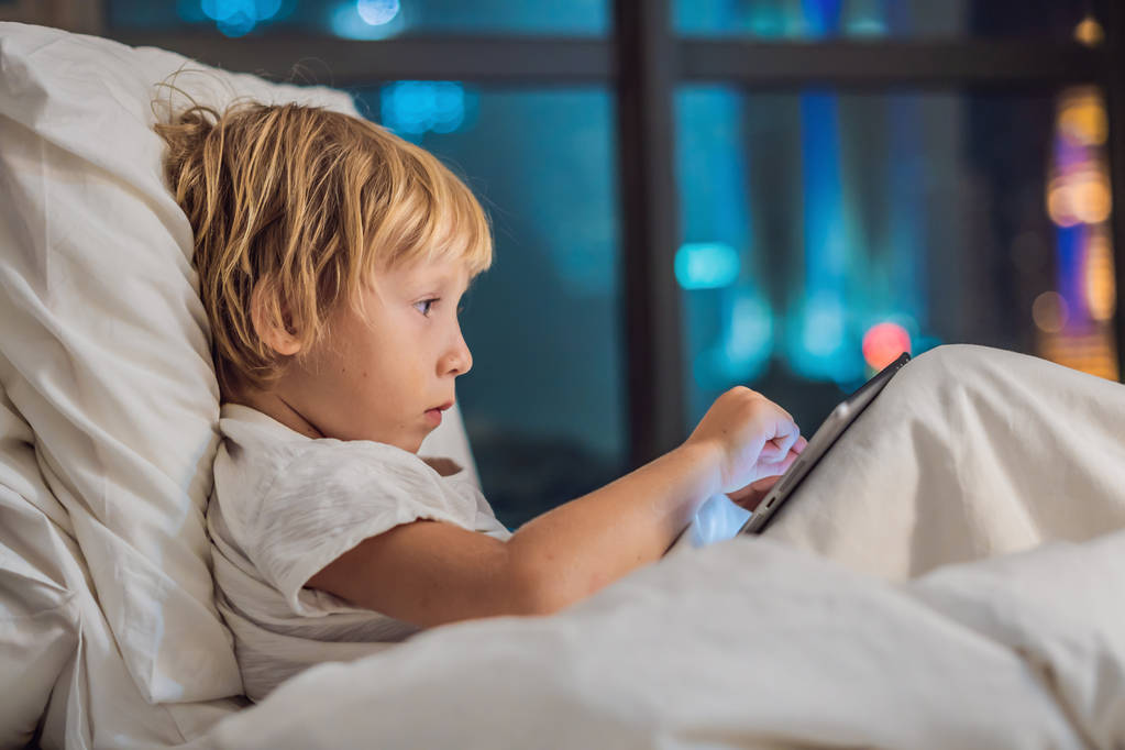 Το αγόρι χρησιμοποιεί το tablet στο κρεβάτι του πριν πάτε για ύπνο σε ένα υπόβαθρο της πόλης το βράδυ. Παιδιά και τεχνολογία έννοιες - Φωτογραφία, εικόνα