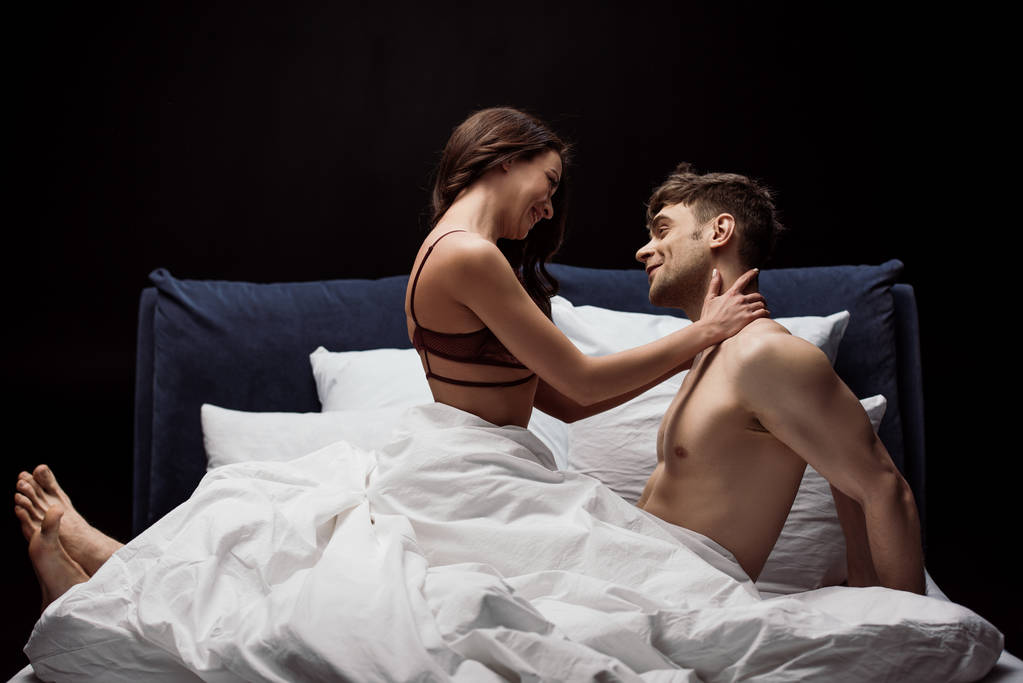 usmívající se žena v sexy prádle všeobjímající muž v posteli izolované na černém pozadí - Fotografie, Obrázek
