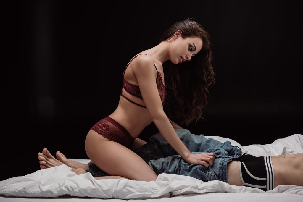 atractiva mujer joven en ropa interior sexy desvestirse hombre en la cama aislado en negro
 - Foto, imagen