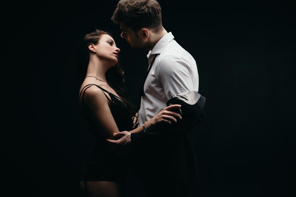 beautiful sexy woman undressing man in formal wear isolated on black - Photo, Image