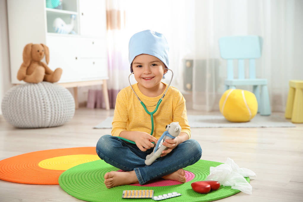 Söpö lapsi kuvittelee itsensä lääkäriksi leikkiessään stetoskoopilla ja lelukarhulla kotona
 - Valokuva, kuva
