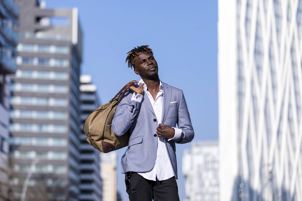Vista frontale nero giovane uomo urbano moda casual in strada. Indossare giacca nera elegante, jeans, tenendo e portando una borsa in una giornata di sole
 - Foto, immagini