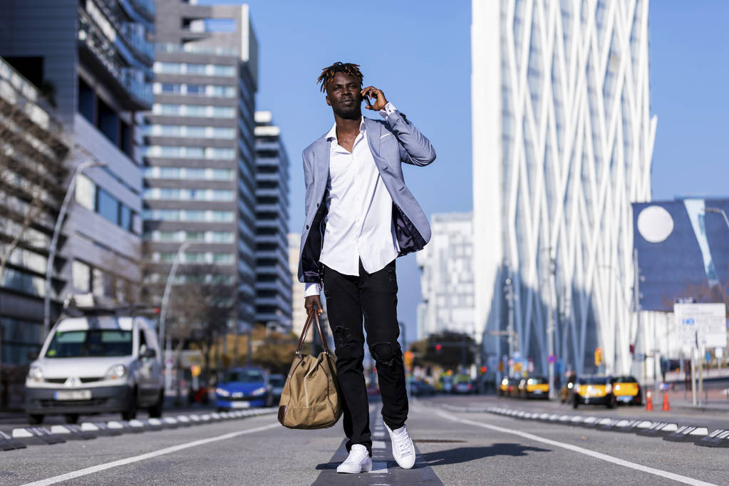Vooraanzicht van een zwarte Afrikaanse jonge man lopen dragen van elegante jas of een tas te houden tijdens het gebruik van de telefoon in de zonnige dag op straat - Foto, afbeelding