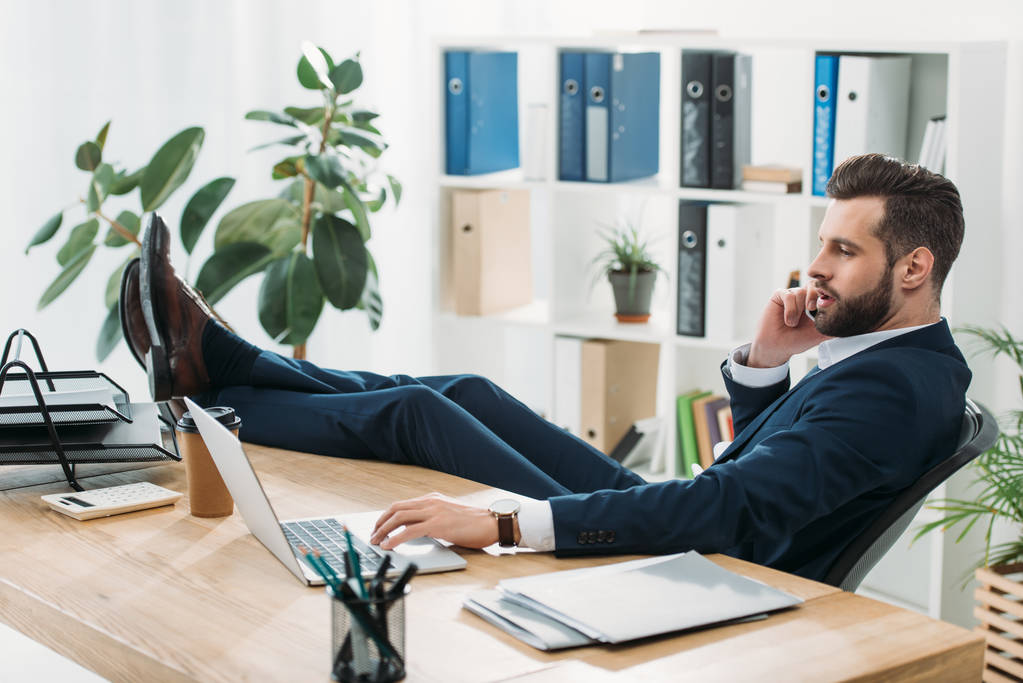 Geschäftsmann im blauen Anzug sitzt mit Beinen am Tisch und telefoniert im Büro mit dem Smartphone - Foto, Bild