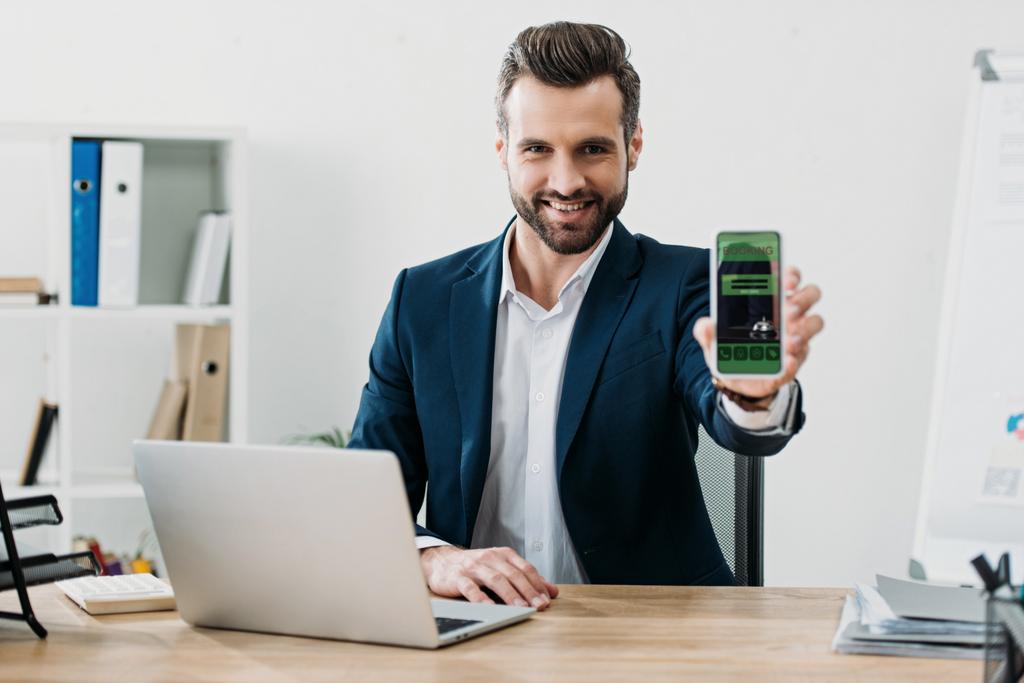 empresário sentado à mesa com laptop e mostrando smartphone com aplicativo de reserva na tela no escritório
 - Foto, Imagem