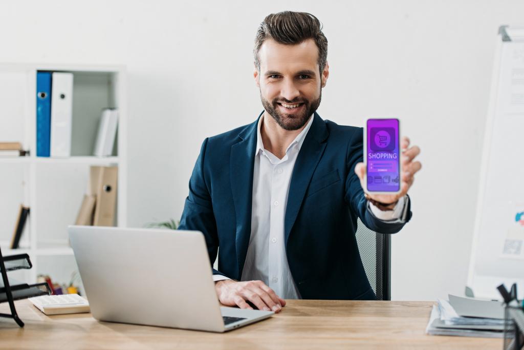 homme d'affaires à table avec ordinateur portable et montrant smartphone avec application shopping à l'écran dans le bureau
 - Photo, image