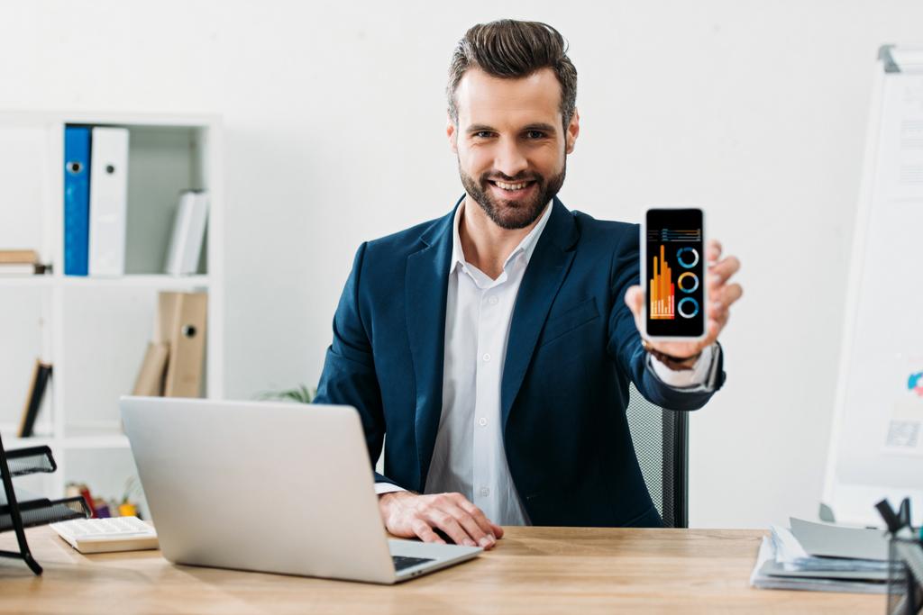 uomo d'affari seduto a tavola con laptop e mostrando smartphone con grafici e grafici app sullo schermo in ufficio
 - Foto, immagini