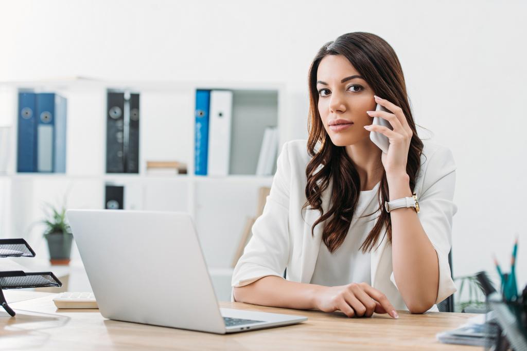 mujer de negocios sentada en la mesa con portátil y hablando en el teléfono inteligente en la oficina
 - Foto, Imagen