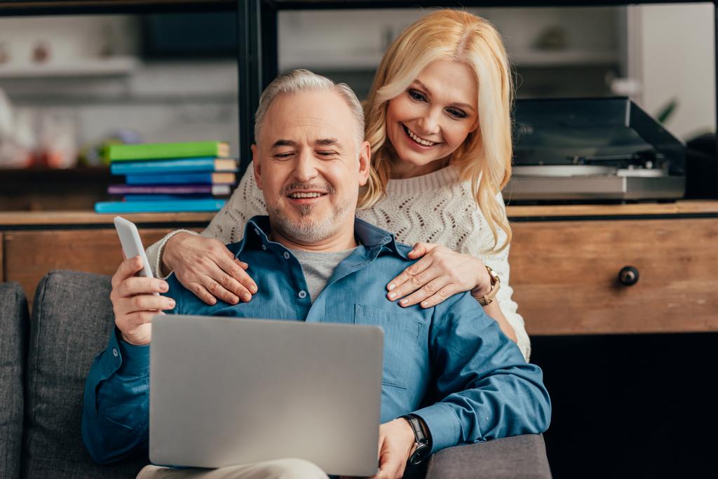 mulher loira abraçando marido sorridente olhando para laptop enquanto segurando smartphone
 - Foto, Imagem
